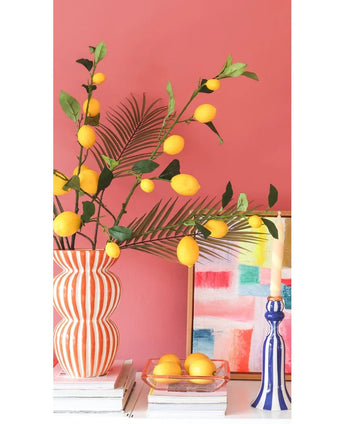 Contra una pared rosa coral hay un jarrón a rayas lleno de ramas de Kunsttak Lemon de 104 cm en amarillo, un cuadro colorido, una pila de libros, un candelabro azul con una vela y una bandeja de limones.