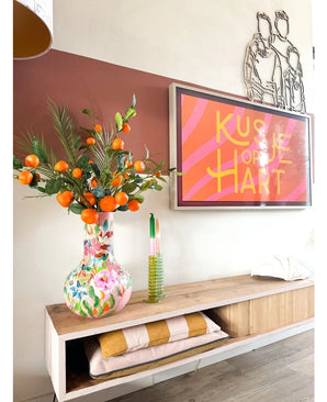 Sobre un estante de madera se encuentra un jarrón colorido que contiene el Art Branch Mandarin 93 cm naranja, junto a una obra de arte enmarcada con rayas rosas que dice “Kissje op je Hart”. También en el estante hay una vela verde y mantas dobladas.