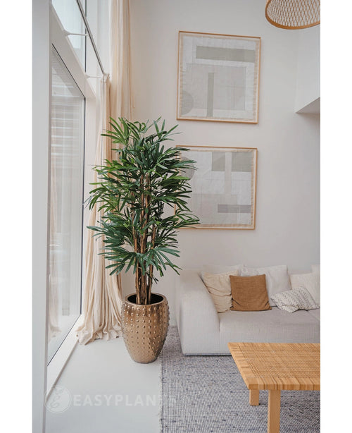 Una Lady Artificial Palm de 180 cm se coloca cerca de un gran ventanal en una sala de estar moderna y luminosa con un sofá blanco, cojines beige, una mesa tejida y arte abstracto. Agrega un estilo minimalista acogedor con sus tonos neutros y el encanto de las plantas artificiales verdes.