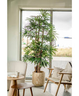 Een 210 cm hoge Lady Kunstpalm staat naast een raam in een lichte, moderne woonkamer met een stoel met kussens, een kleine ronde tafel en een neutrale inrichting.