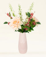 Het Zijden kunstbloemen boeket Morning Whisper - een mix van perzikkleurige dahlia's, witte leeuwenbekjes, roze bessen en groene blaadjes in een lichtroze vaas van keramiek - staat tegen een effen gebroken witte achtergrond.