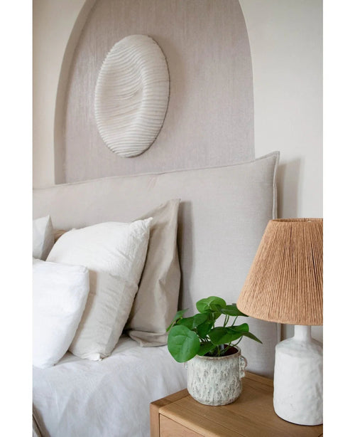 Un acogedor rincón para dormir con una cabecera beige, almohadas blancas y beige, una mesita de noche de madera, una lámpara de cerámica con pantalla tejida, la planta artificial de panqueques de 27 cm y un cuadro blanco en la pared encima de la cama.