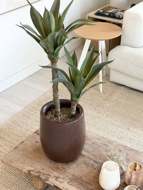Una planta de agave artificial de 76 cm, con dos tallos largos y hojas verdes puntiagudas, se encuentra sobre una mesa de centro de madera en una acogedora sala de estar con un sofá color crema, una mesa auxiliar redonda y una decoración neutra.