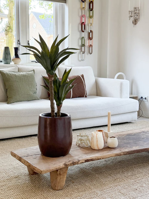 Un salón moderno con un sofá blanco, cojines verdes y marrones, una mesa de centro de madera decorada con velas y la planta de Agave Artificial de 76 cm, y una decoración de pared de malla de cadenas. La luz natural entra por las ventanas detrás del sofá.