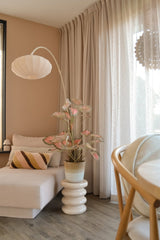 Una acogedora zona de estar con chaise longue beige, cojín de rayas, el Caladium Artificial 90 cm rosa que aporta un toque verde, una lámpara moderna, cortinas claras y suelo de madera. La suave luz natural entra a través de las cortinas transparentes.