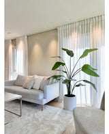 Un salón moderno con un sofá gris claro, cojines blancos, la planta artificial Strelitzia Real Touch confeccionada en un macetero beige de 180 cm, cortinas blancas transparentes, una alfombra beige y una suave luz natural que entra por las ventanas.