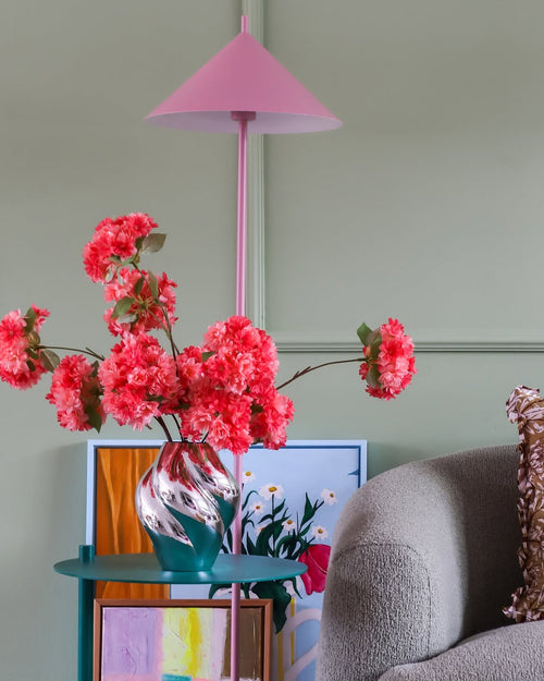 Una lámpara rosa, un jarrón a rayas con el ramo de flores artificiales de seda Coral en flor de cerezo, obras de arte coloridas y una silla gris se apoyan contra una pared verde claro, creando un rincón acogedor y animado.